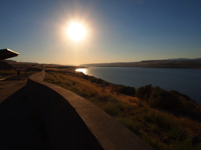 [The sun is a bright white ball with rays extending from it in the sky. A short wall winds into the foreground separating visitors from the vegetation at the river's edge.]